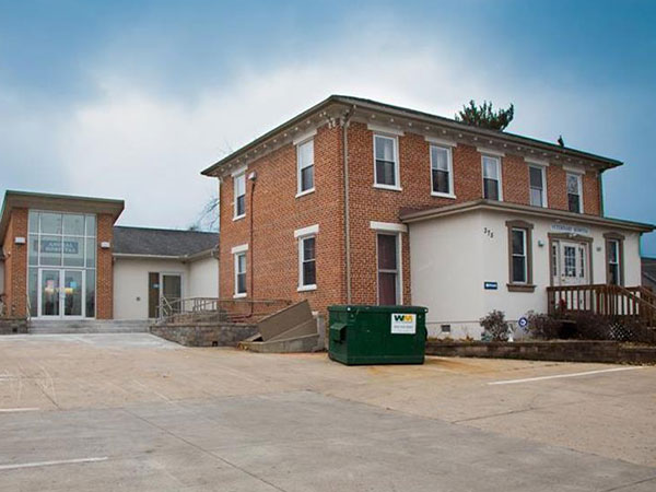 A brick building with white trim and a modern glass extension sits next to a parking lot with a green dumpster in front. The sky is overcast.