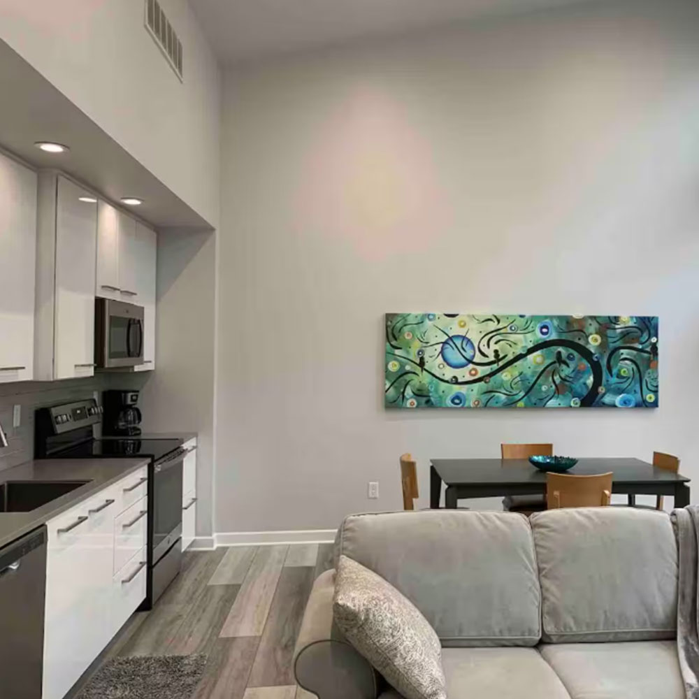 Modern apartment interior with a gray couch in the foreground, a dining table with four chairs and a blue bowl, vibrant abstract wall art, and a kitchen area with white cabinets and stainless steel appliances.