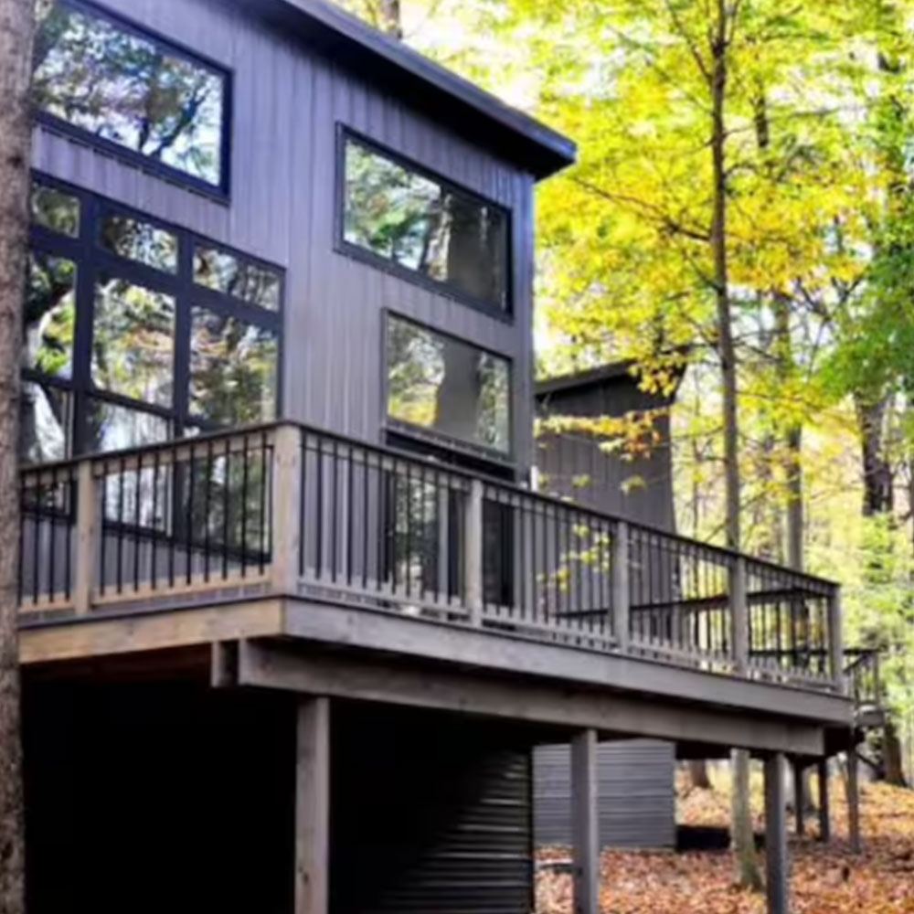 Modern grey cabin with large windows and a spacious wooden deck, situated among tall trees with green and yellow leaves in a forest setting during autumn.